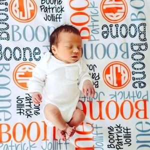 May include: A white baby blanket with a personalized design featuring the names "Boone Patrick Jolliff" in orange and gray text. The blanket is decorated with a repeating pattern of the initials "BJP" in a circle with an orange background. A baby is lying on the blanket.
