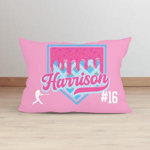 May include: Pink pillow with a baseball diamond design. The diamond is filled with a pink and blue pattern and has a pink donut with sprinkles dripping down the sides. The text "Harrison #16" is on the pillow.