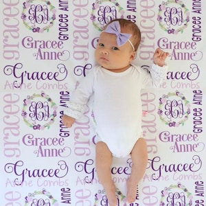 Op de afbeelding: Een wit babydekentje met een paars en roze bloemenpatroon. Het dekentje heeft de naam "Gracee Anne" die door het hele ontwerp heen wordt herhaald. Het dekentje is versierd met een gemonogrammeerd "GA" in een krans van bloemen.