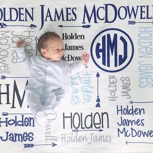 May include: A white baby blanket with a blue and gray pattern. The blanket features the name "Holden James McDowell" repeated throughout the design, along with arrows and a monogram "HMJ".