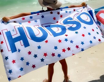 May include: A white beach towel with red and blue stars and the name "HUDSON" printed in blue. A child is holding the towel in front of them, standing on a sandy beach.