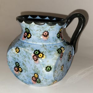 Może przedstawiać: Jasnoniebieski ceramiczny dzbanek z czarnym uchwytem i obramowaniem. Dzbanek jest ozdobiony kolorowymi kwiatowymi wzorami w kolorze różowym, żółtym, zielonym i czarnym. Dzbanek ma teksturowaną powierzchnię.