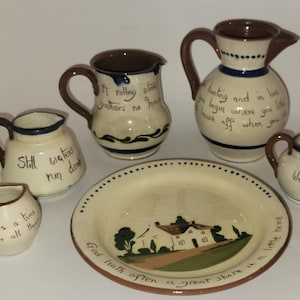 May include: A collection of cream-colored ceramic kitchenware featuring brown handles and rims, including four pitchers and one plate. Each piece is decorated with hand-painted text and a central illustration of a house on the plate.