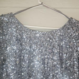 Puede incluir: Una prenda gris plateado de manga larga cubierta de peque&ntilde;as lentejuelas reflectantes. Se ven el escote redondo y las mangas, y la prenda cuelga de una percha blanca sobre un fondo blanco. Las lentejuelas crean un efecto brillante.