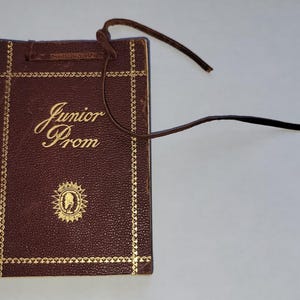 May include: A brown leatherette booklet with gold lettering that reads "Junior Prom." The booklet has a gold border and a small emblem. A brown leather cord is attached to the top of the booklet.