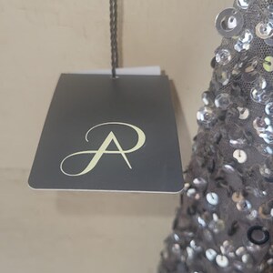 Puede incluir: Una etiqueta gris oscuro con un logotipo estilizado "A" cuelga de un cord&oacute;n negro. La etiqueta est&aacute; sujeta a una prenda gris con lentejuelas. El fondo es una pared de color claro.