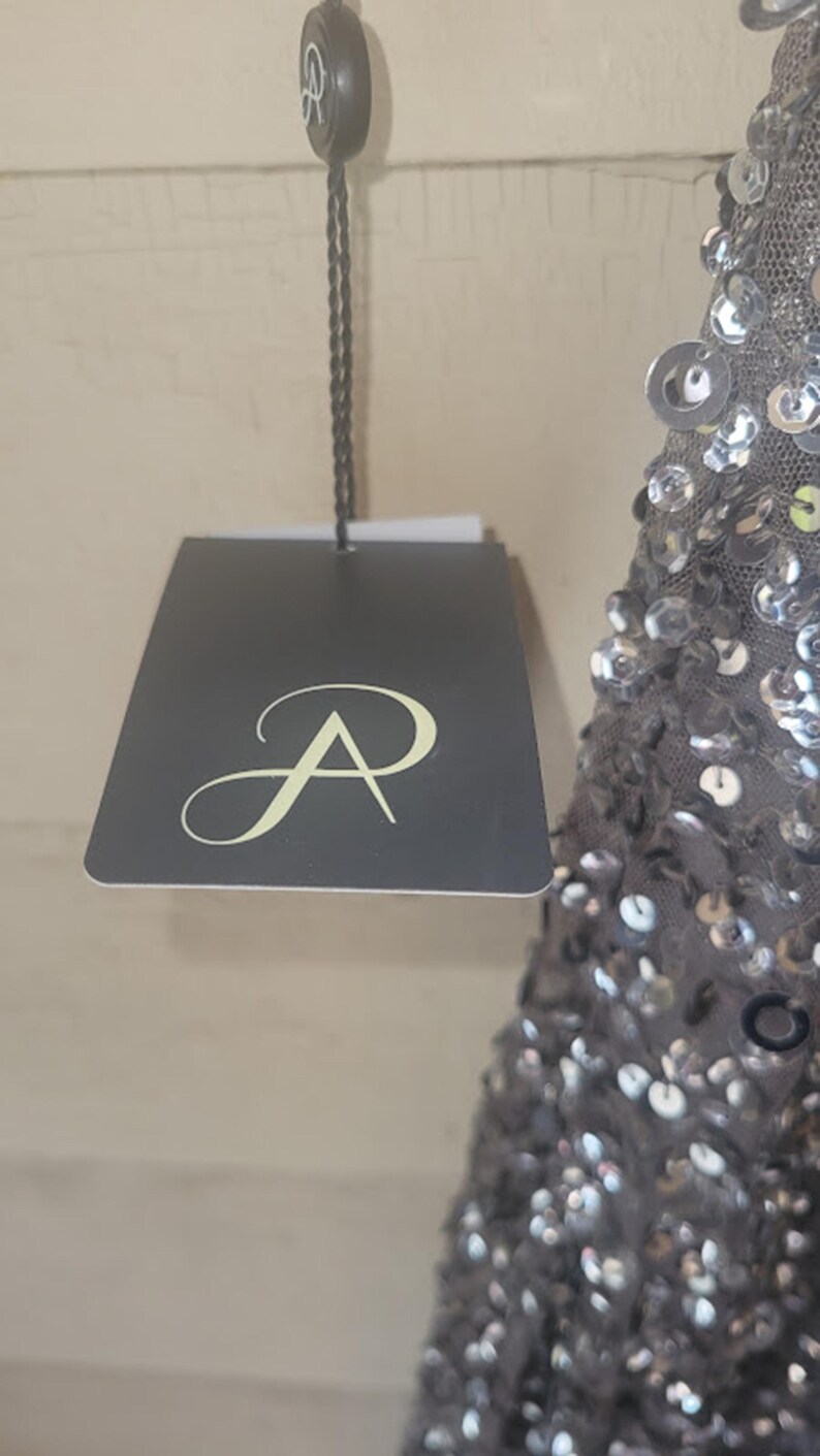 Puede incluir: Una etiqueta gris oscuro con un logotipo estilizado "A" cuelga de un cord&oacute;n negro. La etiqueta est&aacute; sujeta a una prenda gris con lentejuelas. El fondo es una pared de color claro.