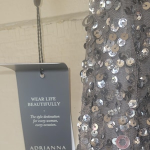 Puede incluir: Un vestido gris con lentejuelas plateadas y el texto "WEAR LIFE BEAUTIFULLY" en una etiqueta. La etiqueta tambi&eacute;n dice "The style destination for every woman, every occasion." Tambi&eacute;n es visible el nombre de la marca "ADRIANNA PAPELL".