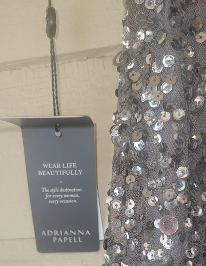 Puede incluir: Un vestido gris con lentejuelas plateadas y el texto "WEAR LIFE BEAUTIFULLY" en una etiqueta. La etiqueta tambi&eacute;n dice "The style destination for every woman, every occasion." Tambi&eacute;n es visible el nombre de la marca "ADRIANNA PAPELL".