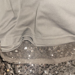Puede incluir: Primer plano de una prenda gris con una superposici&oacute;n de lentejuelas. Las lentejuelas son plateadas y est&aacute;n dispersas sobre una tela transparente. La prenda tiene un forro gris liso y los bordes est&aacute;n cuidadosamente acabados.