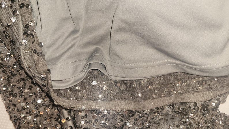 Puede incluir: Primer plano de una prenda gris con una superposici&oacute;n de lentejuelas. Las lentejuelas son plateadas y est&aacute;n dispersas sobre una tela transparente. La prenda tiene un forro gris liso y los bordes est&aacute;n cuidadosamente acabados.
