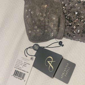 Puede incluir: Una prenda gris con cuentas con una etiqueta de Adrianna Papell. La prenda tiene una secci&oacute;n de lentejuelas y una secci&oacute;n de punto. La etiqueta indica el n&uacute;mero de estilo, el color y la talla 6. La descripci&oacute;n en la etiqueta dice "3/4 SLY BEADED MRMAID GOWN."