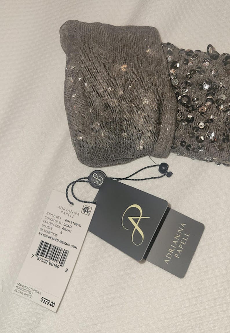 Puede incluir: Una prenda gris con cuentas con una etiqueta de Adrianna Papell. La prenda tiene una secci&oacute;n de lentejuelas y una secci&oacute;n de punto. La etiqueta indica el n&uacute;mero de estilo, el color y la talla 6. La descripci&oacute;n en la etiqueta dice "3/4 SLY BEADED MRMAID GOWN."