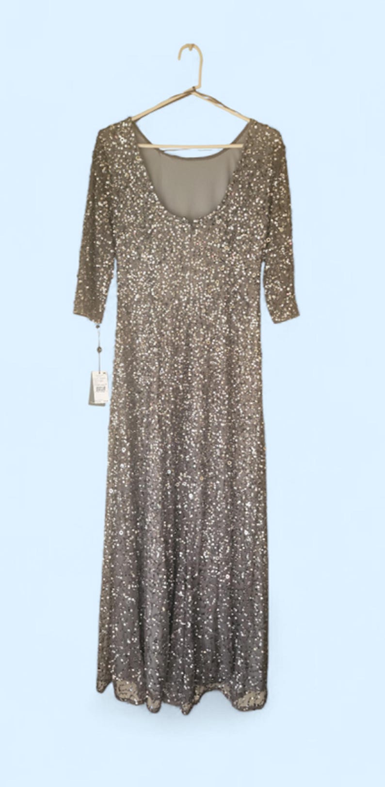 Puede incluir: Un vestido largo de lentejuelas plateadas con mangas tres cuartos cuelga de una percha dorada. El vestido tiene un escote redondo y una silueta entallada. Una etiqueta cuelga del vestido.