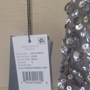 Puede incluir: Un vestido de sirena gris con cuentas con una etiqueta de Adrianna Papell. La etiqueta indica la talla 6, color plomo y n&uacute;mero de estilo 091919970. El vestido presenta lentejuelas plateadas.