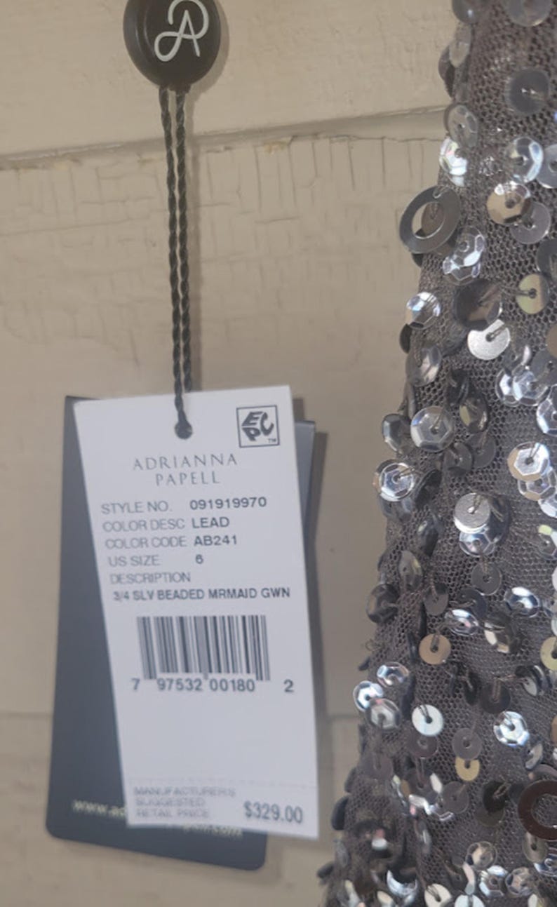 Puede incluir: Un vestido de sirena gris con cuentas con una etiqueta de Adrianna Papell. La etiqueta indica la talla 6, color plomo y n&uacute;mero de estilo 091919970. El vestido presenta lentejuelas plateadas.