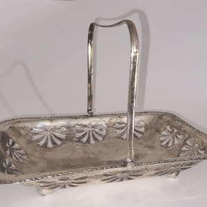 May include: A silver-toned rectangular basket with a handle. The basket features decorative cut-out floral designs and rests on small feet. The handle arches upward, providing a convenient carrying option. The basket is likely used for serving or display.