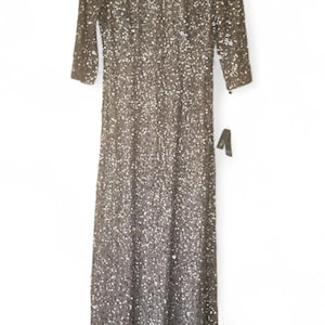 Puede incluir: Vestido largo con mangas tres cuartos, cubierto de lentejuelas brillantes. El vestido tiene un escote redondo y una etiqueta. El vestido es de color neutro, posiblemente plateado o gris.