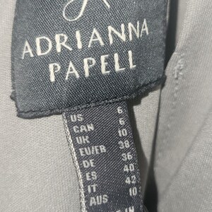 Puede incluir: Primer plano de una etiqueta negra con el nombre de la marca "ADRIANNA PAPELL" en letras blancas. La etiqueta incluye conversiones de tallas para US, CAN, UK, EU/FR, DE, ES, IT y AUS. La etiqueta dice "MADE IN INDIA."
