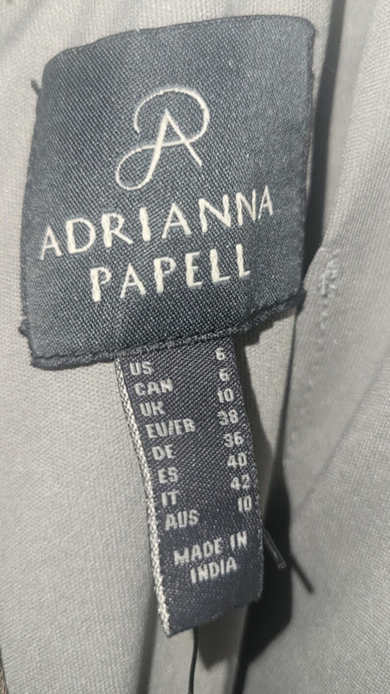 Puede incluir: Primer plano de una etiqueta negra con el nombre de la marca "ADRIANNA PAPELL" en letras blancas. La etiqueta incluye conversiones de tallas para US, CAN, UK, EU/FR, DE, ES, IT y AUS. La etiqueta dice "MADE IN INDIA."