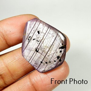 May include: A polished, irregularly shaped gemstone with a lavender hue. The stone features parallel, light-coloured striations and small, dark inclusions. The text "Front Photo" is visible at the bottom of the image.
