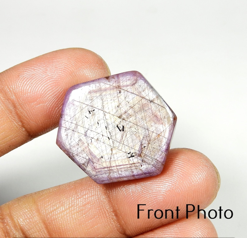 Op de afbeelding: Een zeshoekige, lavendelkleurige edelsteen met interne lijnen en insluitsels. De steen wordt tussen de vingers gehouden, met de tekst "Front Photo" onderaan.