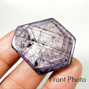 May include: A hexagonal, translucent purple sapphire gemstone featuring visible dark internal inclusions and linear striations across its polished surface. The stone displays a mix of light and deep violet tones with black mineral deposits.