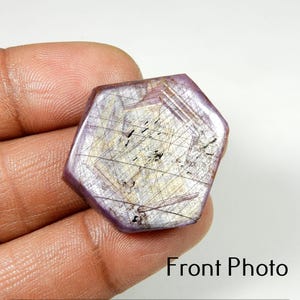 Puede incluir: Una gema hexagonal con una superficie pulida. La piedra presenta una mezcla de tonos morados, grises y dorados con líneas e inclusiones internas visibles. El texto "Front Photo" es visible en la parte inferior.
