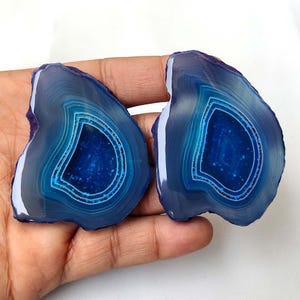 May include: Two polished agate slices featuring concentric bands of deep blue and light blue hues. The centres of these stones display a dark, crystalline texture with small, sparkling inclusions, showcasing natural mineral formations.