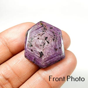 Puede incluir: Una gema facetada, hexagonal, de color púrpura intenso, con patrones internos en forma de estrella e inclusiones oscuras. La gema se sostiene en una mano, con el texto "Front Photo" visible.
