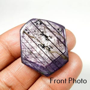 Peut inclure: Une pierre précieuse hexagonale facettée, de couleur violet profond. La pierre présente un réseau d'inclusions linéaires et des motifs sombres et irréguliers. Le texte "Front Photo" est visible en bas de l'image. La pierre est tenue dans une main.