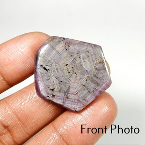 May include: A flat, polished, hexagonal-shaped gemstone with a translucent light purple and lavender hue. The surface displays visible internal inclusions, dark mineral flecks, and a geometric pattern of intersecting lines throughout the stone.