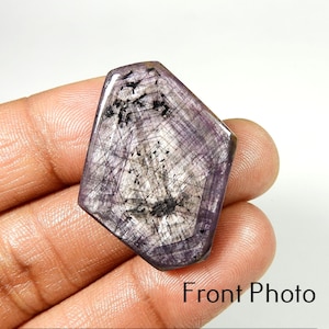 Peut inclure: Une pierre précieuse facettée de forme irrégulière unique. La pierre présente un mélange de teintes violettes, grises et noires, avec des motifs internes complexes. La surface est polie et réfléchissante, mettant en valeur la beauté naturelle du minéral. Le texte "Front Photo" est visible.