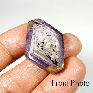 May include: A polished, diamond-shaped gemstone featuring a translucent purple and white color palette with visible internal inclusions and dark black mineral streaks. The stone has a smooth, faceted surface and a distinct geometric shape.