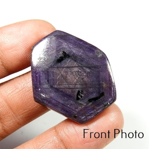 May include: A faceted, hexagonal-shaped purple gemstone with a polished surface, featuring visible internal geometric patterns and dark inclusions throughout the stone. The gem displays a deep violet hue with light reflecting off its flat facets.