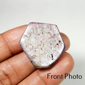 Puede incluir: Una gema hexagonal de color púrpura claro con una red de finas líneas e inclusiones oscuras. La piedra se sostiene en una mano, con el texto "Front Photo" visible en la parte inferior.