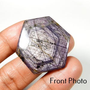 May include: A hexagonal polished gemstone featuring a deep purple and violet hue with visible internal striations, natural inclusions, and a geometric pattern. The surface displays a smooth, reflective finish with dark mineral veins throughout.