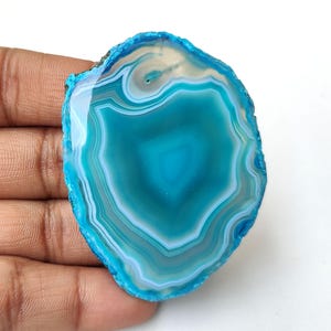 May include: A polished, oval-shaped agate stone slice. The stone features concentric bands of teal, turquoise, and white, with a vibrant blue outer edge. The natural gemstone has a smooth, glossy surface and a unique, organic pattern.
