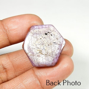 Op de afbeelding: Een zeshoekige, gepolijste edelsteen met een lichtpaarse tint en interne lineaire insluitsels. De steen wordt in een hand gehouden, met de tekst "Back Photo" onderaan zichtbaar. Het oppervlak van de edelsteen heeft een subtiele, reflecterende glans.
