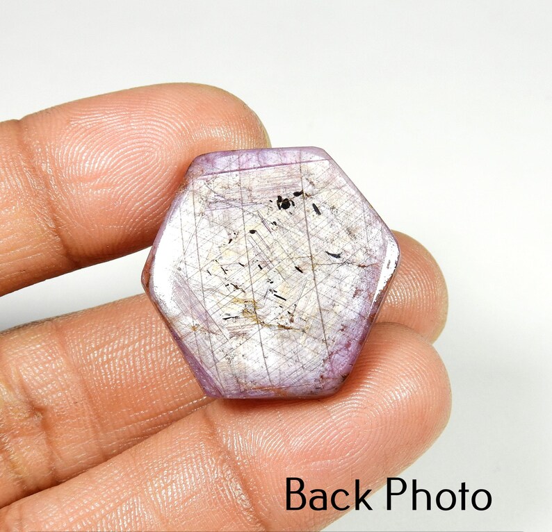 Op de afbeelding: Een zeshoekige, gepolijste edelsteen met een lichtpaarse tint en interne lineaire insluitsels. De steen wordt in een hand gehouden, met de tekst "Back Photo" onderaan zichtbaar. Het oppervlak van de edelsteen heeft een subtiele, reflecterende glans.
