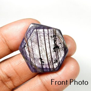 Peut inclure: Une pierre précieuse hexagonale facettée de couleur violet foncé. La pierre présente une structure interne complexe avec des lignes et des inclusions visibles, et une surface polie. Le texte "Front Photo" est visible.