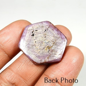 Op de afbeelding: Een gefacetteerde, zeshoekige edelsteen met een lichtpaarse tint en interne adering. De steen heeft een gepolijst oppervlak en wordt in een hand gehouden. De tekst "Back Photo" is zichtbaar onderaan de afbeelding. Close-up van de edelsteen.