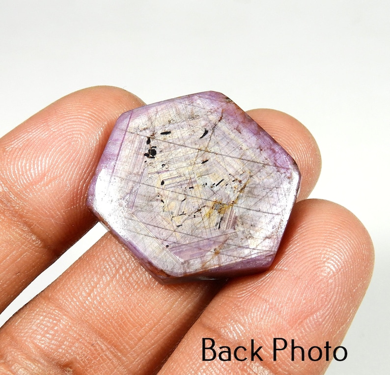 Op de afbeelding: Een gefacetteerde, zeshoekige edelsteen met een lichtpaarse tint en interne adering. De steen heeft een gepolijst oppervlak en wordt in een hand gehouden. De tekst "Back Photo" is zichtbaar onderaan de afbeelding. Close-up van de edelsteen.