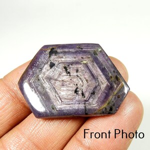 Op de afbeelding: Een zeshoekige, gepolijste edelsteen met een dieppaarse tint en ingewikkelde interne patronen. De steen heeft een glanzend oppervlak en wordt in een hand gehouden. De tekst "Front Photo" is rechtsonder zichtbaar.