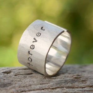 Puede incluir: Un anillo de plata con la palabra "forever" grabada. El anillo está sobre una pieza de madera desgastada.