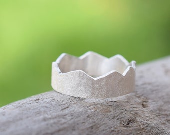 May include: A silver ring with a jagged, mountain-like design. The ring is sitting on a piece of weathered wood.
