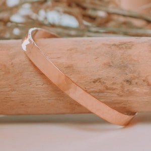 May include: A simple gold bangle bracelet with a hammered finish. The bracelet is resting on a piece of wood.