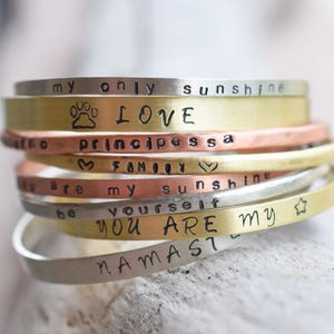 May include: A stack of silver, gold, and copper bracelets with various engraved messages, including "LOVE", "principessa", "FAMILY", "sunshine", "be yourself", "YOU ARE MY", and "NAMASTE".