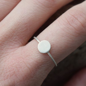 Könnte beinhalten: Ein minimalistischer Silberring mit einem kleinen, kreisförmigen Scheibenmotiv. Der Ring hat ein schmales Band, und die Scheibe hat eine matte Oberfläche. Der Ring wird an einem Finger getragen, was sein schlichtes Design hervorhebt.