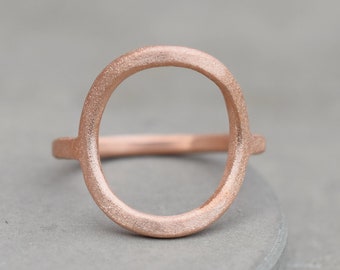 May include: A simple, minimalist ring made of rose gold metal. The ring is a thin band with a large, circular opening in the center.